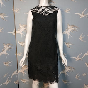 Könnte beinhalten: Schwarzes Spitzenkleid ohne Ärmel auf einer Schaufensterpuppe. Das Kleid hat einen hohen Ausschnitt mit Spitzendetail und einen knielangen Saum. Der Hintergrund ist eine hellblaue Wand mit einem Muster aus weißen Vögeln.