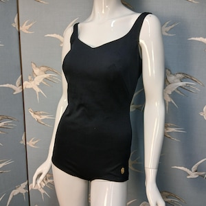 Op de afbeelding: Een zwart badpak met een V-hals en dunne bandjes, gepresenteerd op een mannequin. Een klein gouden embleem is zichtbaar aan de linkerkant. De achtergrond is een blauw behang met witte vogelillustraties. Het is een badpak uit één stuk.