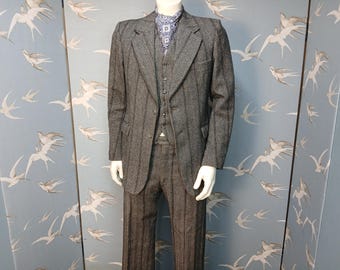 May include: A grey three-piece suit with vertical stripes, comprising a blazer, waistcoat, and trousers, displayed on a mannequin. A patterned scarf is visible. The background is a blue wallpaper with a bird pattern. The suit is a classic style.