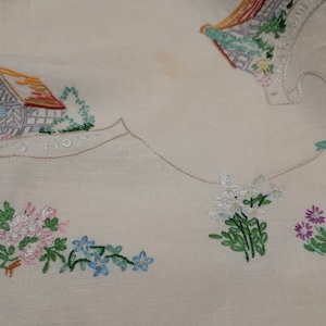 May include: White linen tablecloth with embroidered houses and flowers. The text "Garden's Home" is embroidered on the tablecloth.