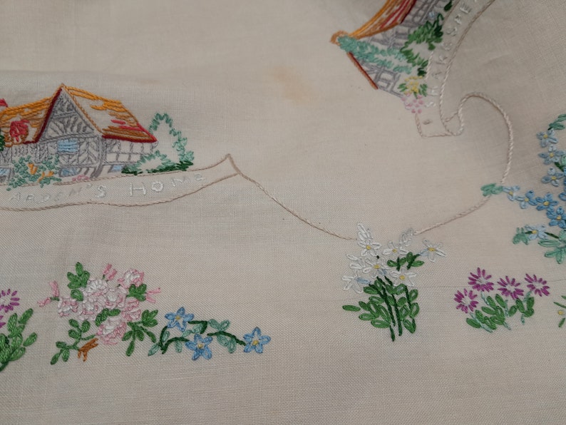 May include: White linen tablecloth with embroidered houses and flowers. The text "Garden's Home" is embroidered on the tablecloth.