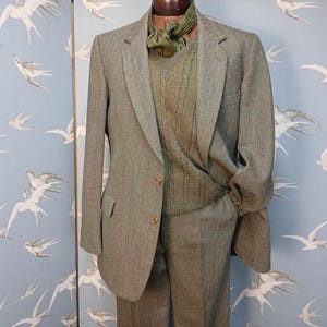 May include: A gray tweed suit, including a blazer and trousers, displayed on a mannequin. The blazer has two buttons and a pocket. A green cable-knit sweater and a green scarf are worn underneath. The background features a blue wallpaper with white bird illustrations.