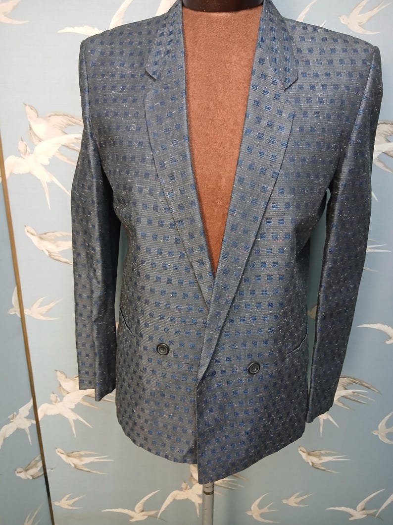 May include: A blue blazer with a subtle checkered pattern. The double-breasted jacket has two rows of buttons and notched lapels. The fabric has a slight sheen, and the blazer is displayed on a mannequin.