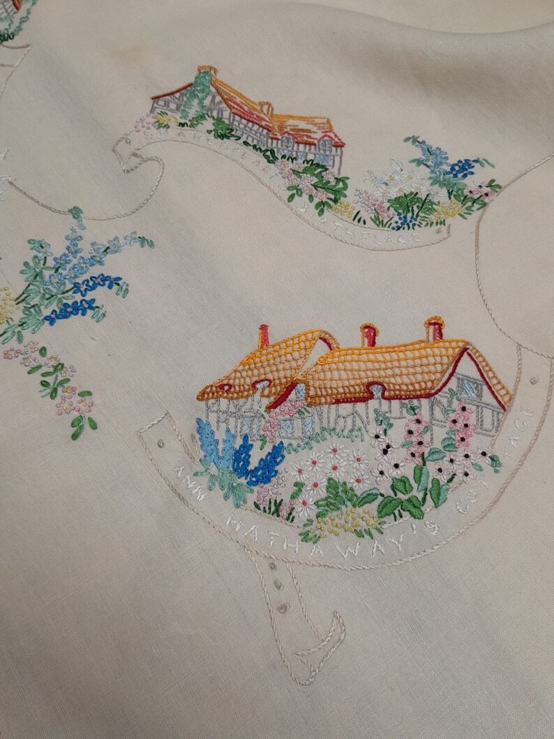 May include: White linen tablecloth with embroidered images of houses and flowers. The text "Ann Hathaway's Way is the Best Way" is embroidered around the bottom of the houses.