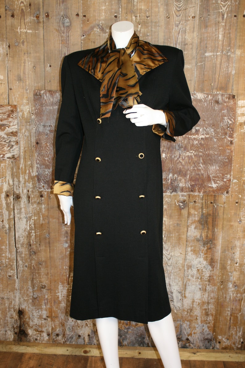 K&ouml;nnte beinhalten: Ein schwarzes, zweireihiges Kleid mit einem goldenen Schal-Kragen mit Tigermuster und goldenen Kn&ouml;pfen. Das Kleid hat lange &Auml;rmel mit goldenen Manschetten mit Tigermuster.