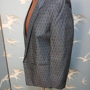 May include: A grey blazer with a blue square pattern is displayed on a mannequin. The blazer has a two-button closure and a notched lapel. The fabric has a subtle sheen, and the blazer appears to be tailored. The background features a light blue wallpaper with white bird illustrations.