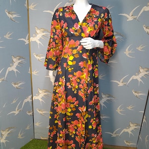May include: A long, flowing maxi dress with a V-neckline and bell sleeves. The dress is made of a sheer fabric with a floral print in shades of pink, yellow, and brown on a dark blue background.