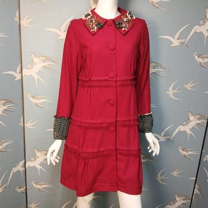 Könnte beinhalten: Ein roter Mantel mit dekorativem Kragen und Knopfleiste. Der Kragen ist mit Perlen und Stickereien verziert. Die Ärmel haben schwarz-weiße Polka-Dot-Manschetten. Der Mantel hat ein gestuftes Rockdesign.