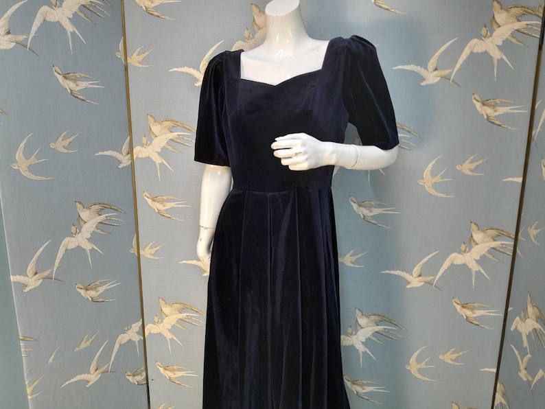 May include: A navy blue velvet dress with a square neckline and short puff sleeves. The dress has a fitted bodice and a full skirt.