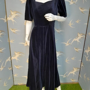 May include: A long, navy blue velvet dress with a square neckline and short puff sleeves. The dress has a fitted bodice and a flowing skirt.