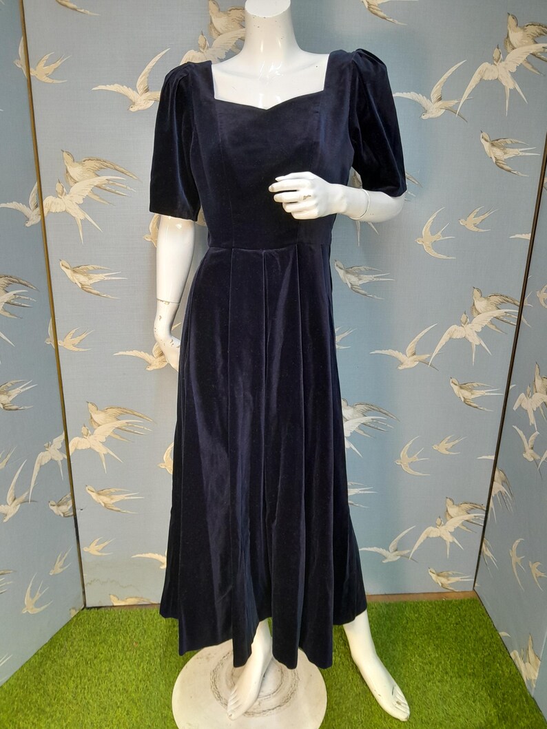 May include: A long, navy blue velvet dress with a square neckline and short puff sleeves. The dress has a fitted bodice and a flowing skirt.