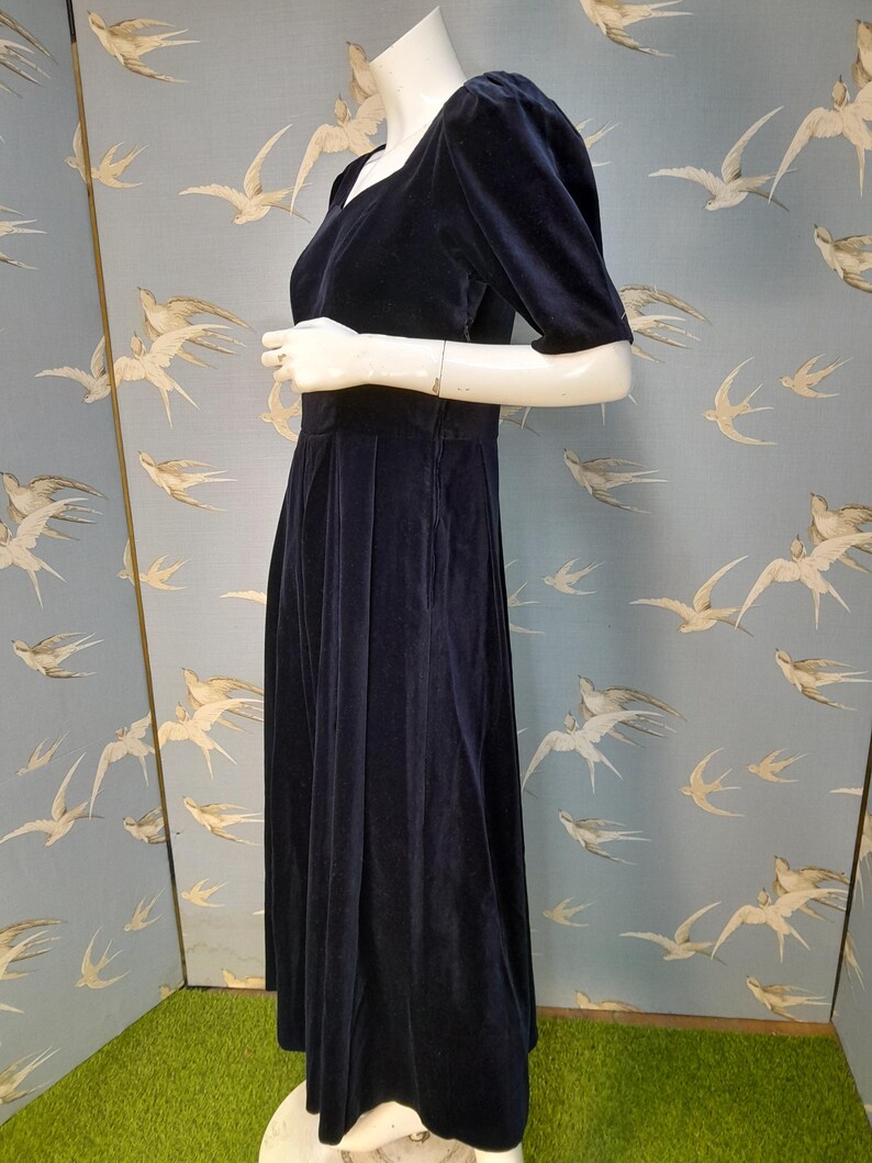 May include: A navy blue velvet dress with a square neckline and short puff sleeves. The dress has a fitted bodice and a flowing skirt.