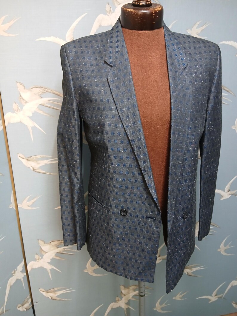 May include: A double-breasted blazer in a blue and silver checked pattern. The jacket is displayed on a mannequin, highlighting its tailored fit and classic design. The blazer features two rows of buttons and a notched lapel.