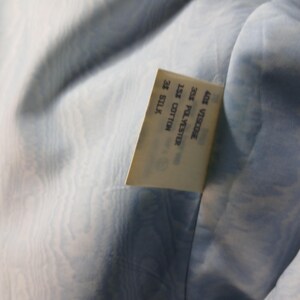 May include: Close-up of a pale blue fabric with a subtle, swirling pattern. A rectangular tag is sewn into the fabric, displaying the composition: 40% Viscose, 25% Polyester, 15% Cotton, and 20% Silk.