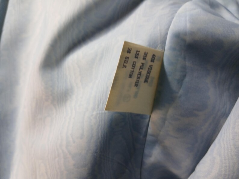 May include: Close-up of a pale blue fabric with a subtle, swirling pattern. A rectangular tag is sewn into the fabric, displaying the composition: 40% Viscose, 25% Polyester, 15% Cotton, and 20% Silk.