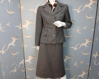May include: A gray skirt suit with a matching jacket. The jacket has a notched collar, three buttons, and decorative buckles. The skirt is knee-length. A patterned scarf is worn under the jacket collar.