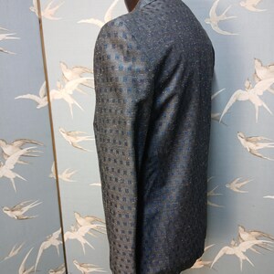 May include: A dark blue blazer with a subtle square pattern and a two-button closure. The jacket is displayed on a mannequin, showcasing its tailored fit and textured fabric. The blazer's design incorporates a mix of dark blue and lighter blue tones.