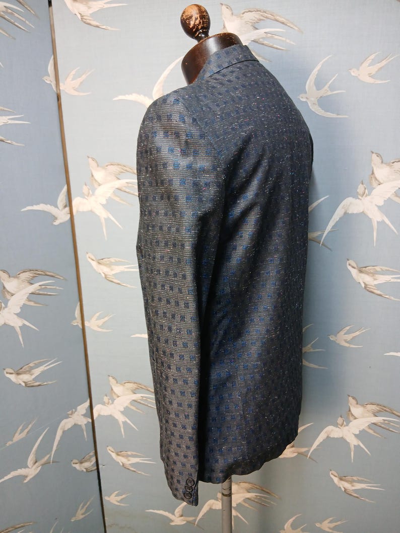 May include: A dark blue blazer with a subtle square pattern and a two-button closure. The jacket is displayed on a mannequin, showcasing its tailored fit and textured fabric. The blazer's design incorporates a mix of dark blue and lighter blue tones.