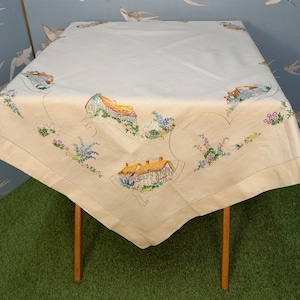 May include: A white tablecloth with embroidered images of houses and flowers. The tablecloth is laid on a wooden table with a green surface underneath.