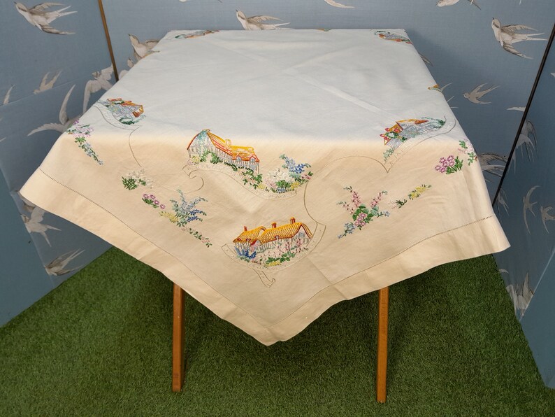 May include: A white tablecloth with embroidered images of houses and flowers. The tablecloth is laid on a wooden table with a green surface underneath.