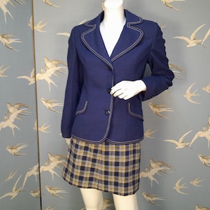 May include: A navy blue blazer with a white trim and a plaid skirt. The blazer has two buttons and a pocket on each side. The skirt is a plaid pattern with yellow, blue, and white.