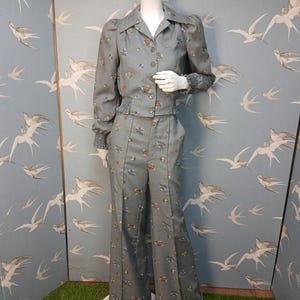 Puede incluir: Un conjunto de dos piezas gris con un estampado floral. El conjunto incluye una camisa abotonada con cuello y mangas largas, y pantalones de pernera ancha. El atuendo se exhibe en un maniquí. El fondo presenta una pared azul con un estampado de pájaros.