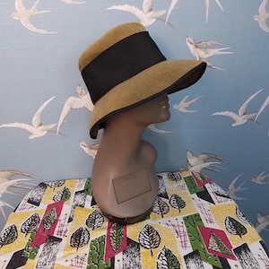 Op de afbeelding: Een vintage hoed met een brede rand en een zwarte band. De hoed wordt getoond op een mannequinhoofd. De kleur van de hoed is lichtbruin. De achtergrond is een blauwe muur met witte vogelillustraties.