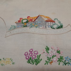 May include: Embroidered white linen tablecloth with a house and flowers. The house is brown with a red roof and green trees. The text "Mary Arden's Home" is embroidered in a banner above the house.