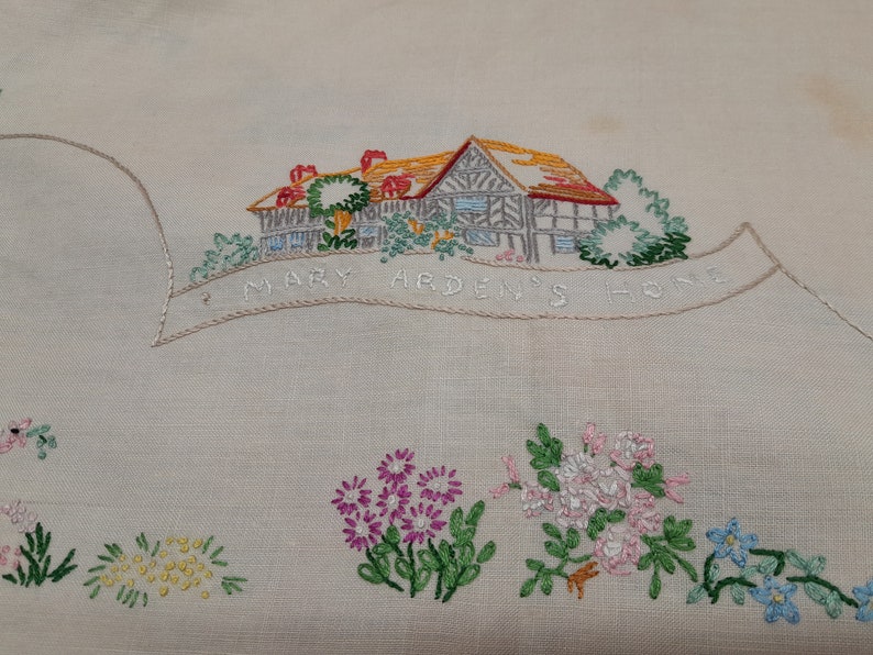 May include: Embroidered white linen tablecloth with a house and flowers. The house is brown with a red roof and green trees. The text "Mary Arden's Home" is embroidered in a banner above the house.