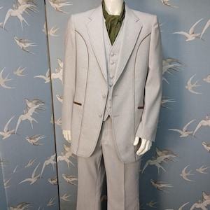 May include: A light grey three-piece suit with brown trim and matching trousers. The suit comprises a jacket, waistcoat, and trousers. The mannequin is wearing a green scarf. The background is a blue wall with a bird pattern.