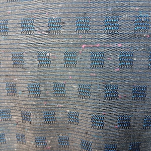 May include: Close-up of a textured fabric with a dark grey base. The fabric features a repeating pattern of small, square designs in shades of blue and black, with subtle pink flecks throughout. The weave is visible.