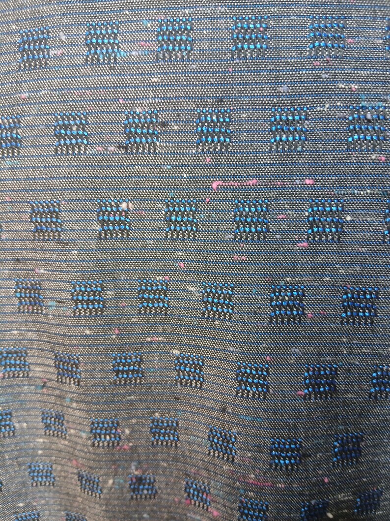 May include: Close-up of a textured fabric with a dark grey base. The fabric features a repeating pattern of small, square designs in shades of blue and black, with subtle pink flecks throughout. The weave is visible.