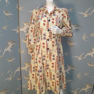 Może przedstawiać: Długa sukienka z długimi rękawami, zapinana na guziki z przodu, w geometryczny wzór w odcieniach czerwieni, błękitu i bieli na kremowym tle.