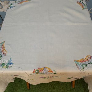 May include: White linen tablecloth with embroidered cottage scenes in shades of yellow, red, green, and blue.