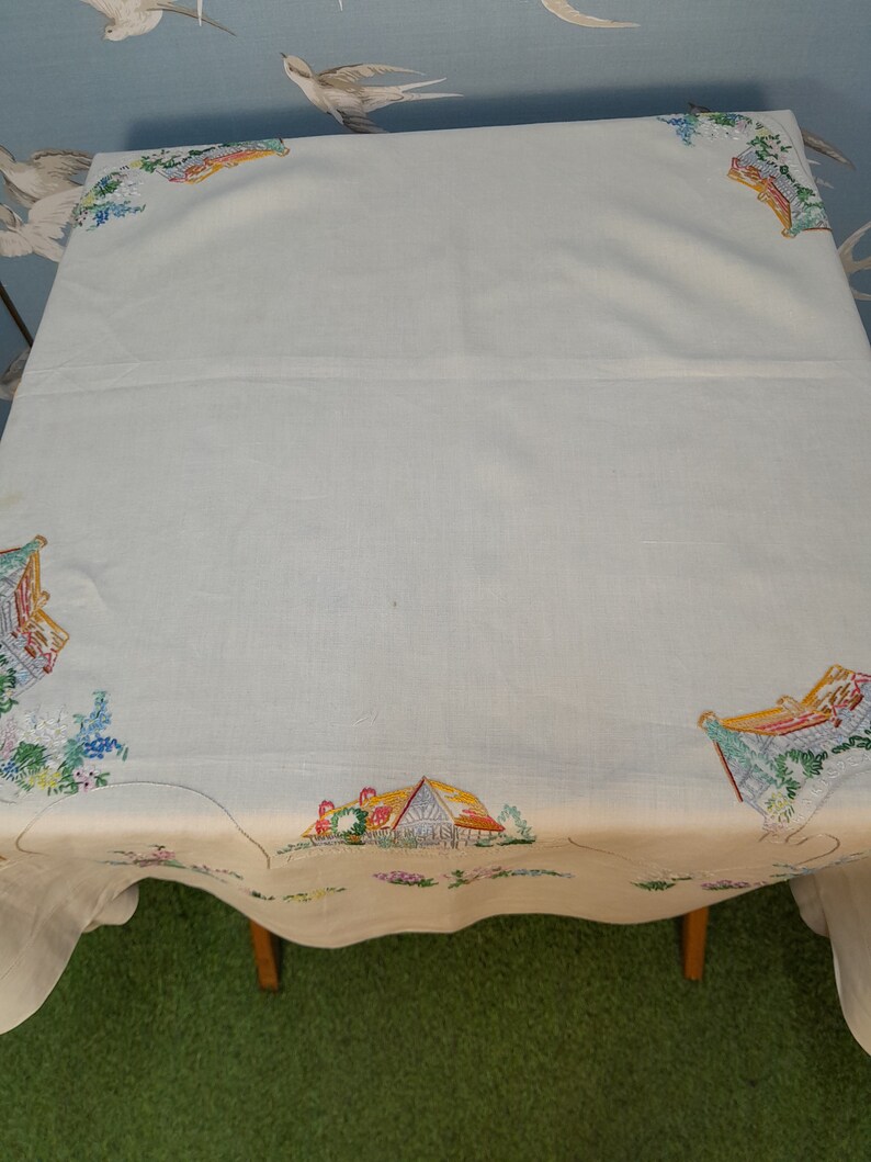 May include: White linen tablecloth with embroidered cottage scenes in shades of yellow, red, green, and blue.