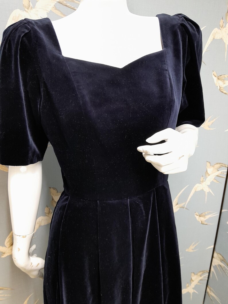 May include: A black velvet dress with a square neckline and short puff sleeves. The dress has a fitted bodice and a full skirt.