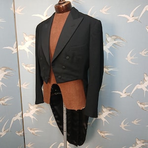 Peut inclure: Un frac noir sur un mannequin. La veste présente un revers cranté, des boutons à double boutonnage et une finition satinée. Le mannequin est présenté sur un fond bleu clair avec un motif d'oiseaux.