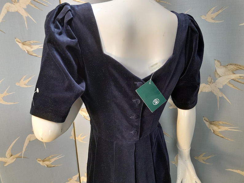 May include: A navy blue velvet dress with a square neckline and puff sleeves. The dress has a button closure down the front and a green tag attached to the side.