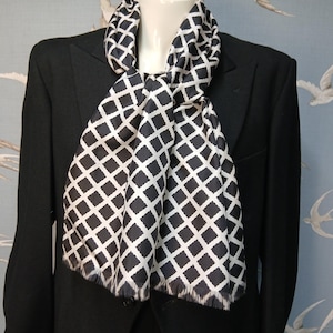 May include: A black scarf featuring a white grid pattern draped over a black blazer. The scarf has a smooth texture and a fringed edge at the bottom. The white lines form a repeating diamond or square lattice design across the dark fabric surface.
