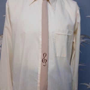 Puede incluir: Una camisa de manga larga color crema con bolsillo en el pecho, combinada con una corbata de color tostado claro. La corbata está decorada con un símbolo de clave de sol en color marrón oscuro colocado verticalmente hacia la parte inferior.