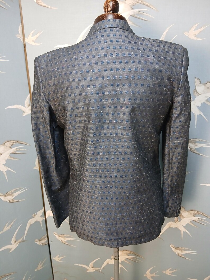 May include: A dark blue blazer with a textured, square pattern. The back of the jacket is visible, showcasing the tailored fit and the subtle sheen of the fabric. The blazer is displayed on a mannequin.