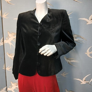 May include: A black velvet blazer with a V-neck and three buttons. The jacket is displayed on a mannequin with red pants. The background features a blue wallpaper with white bird illustrations. The blazer has long sleeves.