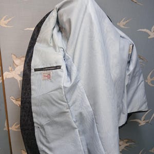 May include: A dark grey blazer with a light blue interior lining. The blazer has an inside pocket and a label. The lining has a subtle pattern. The blazer is shown against a light blue background with a bird pattern.