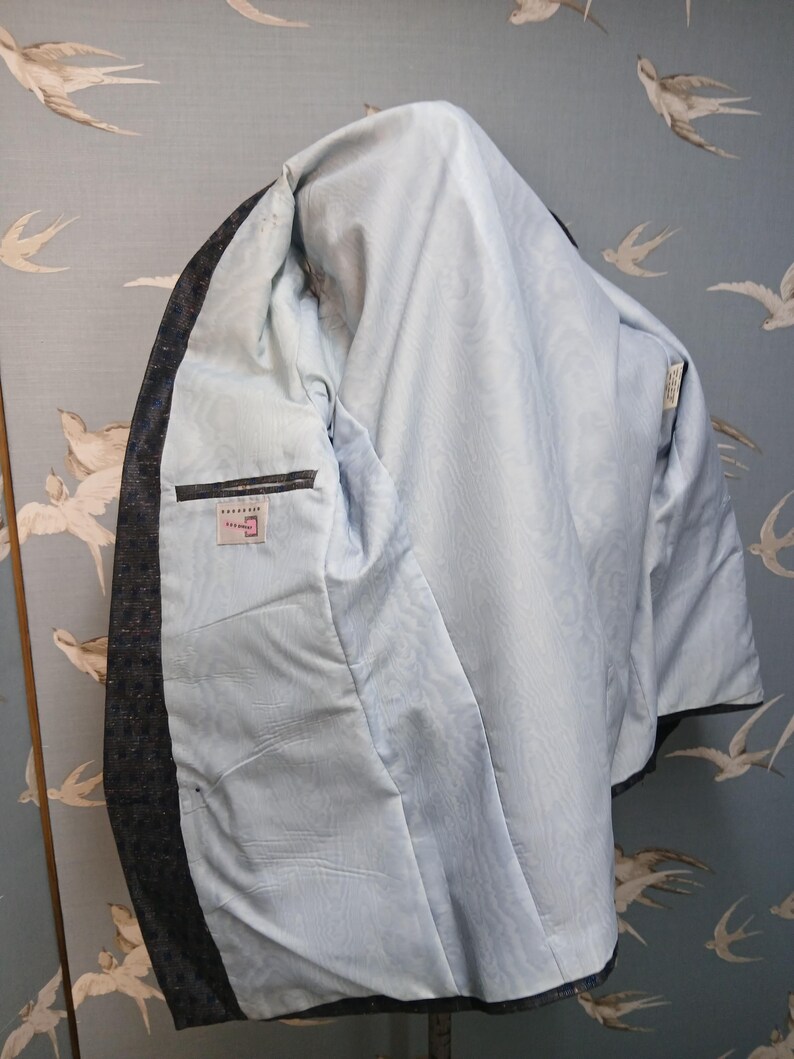 May include: A dark grey blazer with a light blue interior lining. The blazer has an inside pocket and a label. The lining has a subtle pattern. The blazer is shown against a light blue background with a bird pattern.