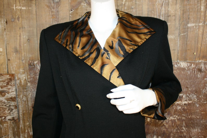 K&ouml;nnte beinhalten: Ein schwarzer Blazer mit einem gro&szlig;en, breiten Kragen, der mit einem braunen und schwarzen Tiermusterstoff gef&uuml;ttert ist. Der Blazer hat einen zweireihigen Verschluss mit einem goldenen Knopf.