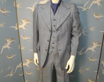 May include: A three-piece grey plaid suit, comprising a jacket, waistcoat, and trousers, displayed on a mannequin. The jacket has a notched lapel and two front pockets. A blue cravat is worn under the jacket. The suit is a classic style.