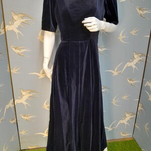 May include: A long, navy blue velvet dress with a square neckline and short puff sleeves. The dress has a fitted bodice and a flowing skirt.