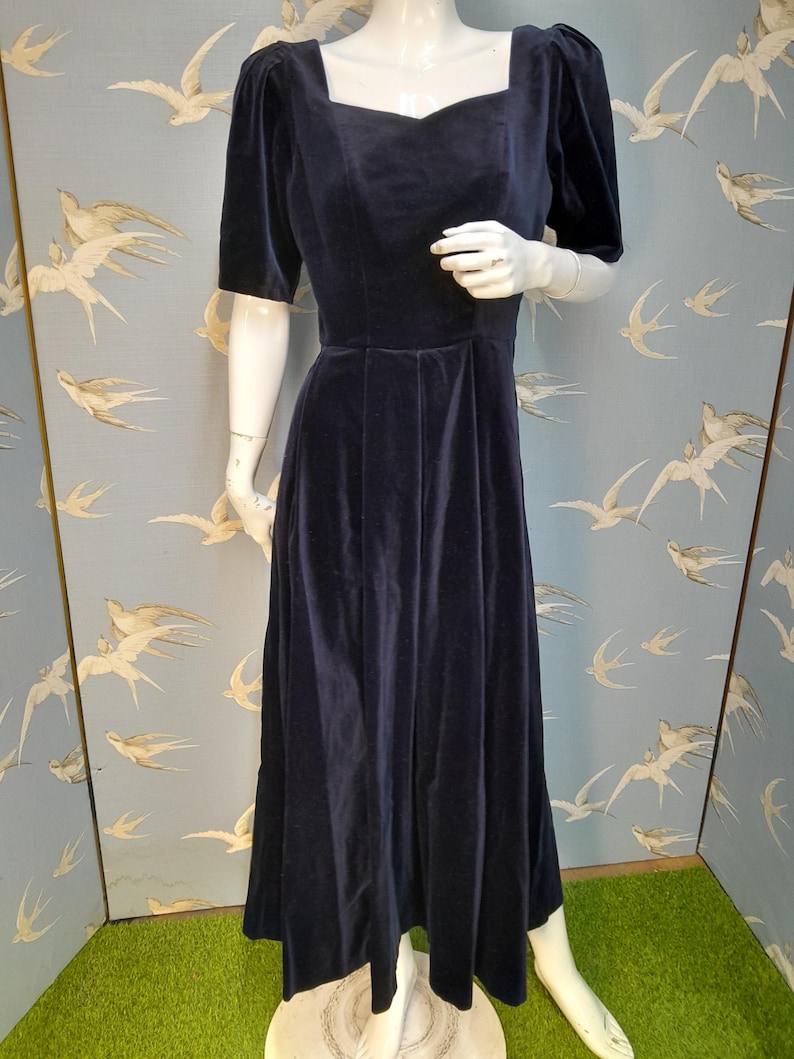 May include: A long, navy blue velvet dress with a square neckline and short puff sleeves. The dress has a fitted bodice and a flowing skirt.