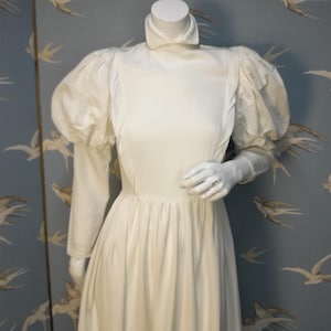 May include: A white vintage wedding dress with a high neck and long sleeves. The dress has a full skirt and puffed sleeves.