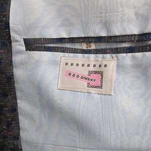 May include: Close-up of a jacket's interior, showing a light blue patterned lining. The jacket's outer fabric is dark and textured, with blue and pink flecks. A tag reads "DDD DIREKT" and the size is 36.